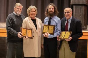 four people stand with plaques in high school auditorium